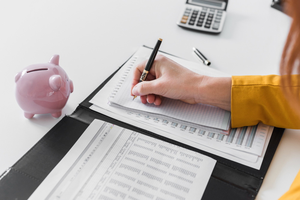 Pessoa organiza anotações e documentos financeiros ao lado de um cofrinho e uma calculadora.